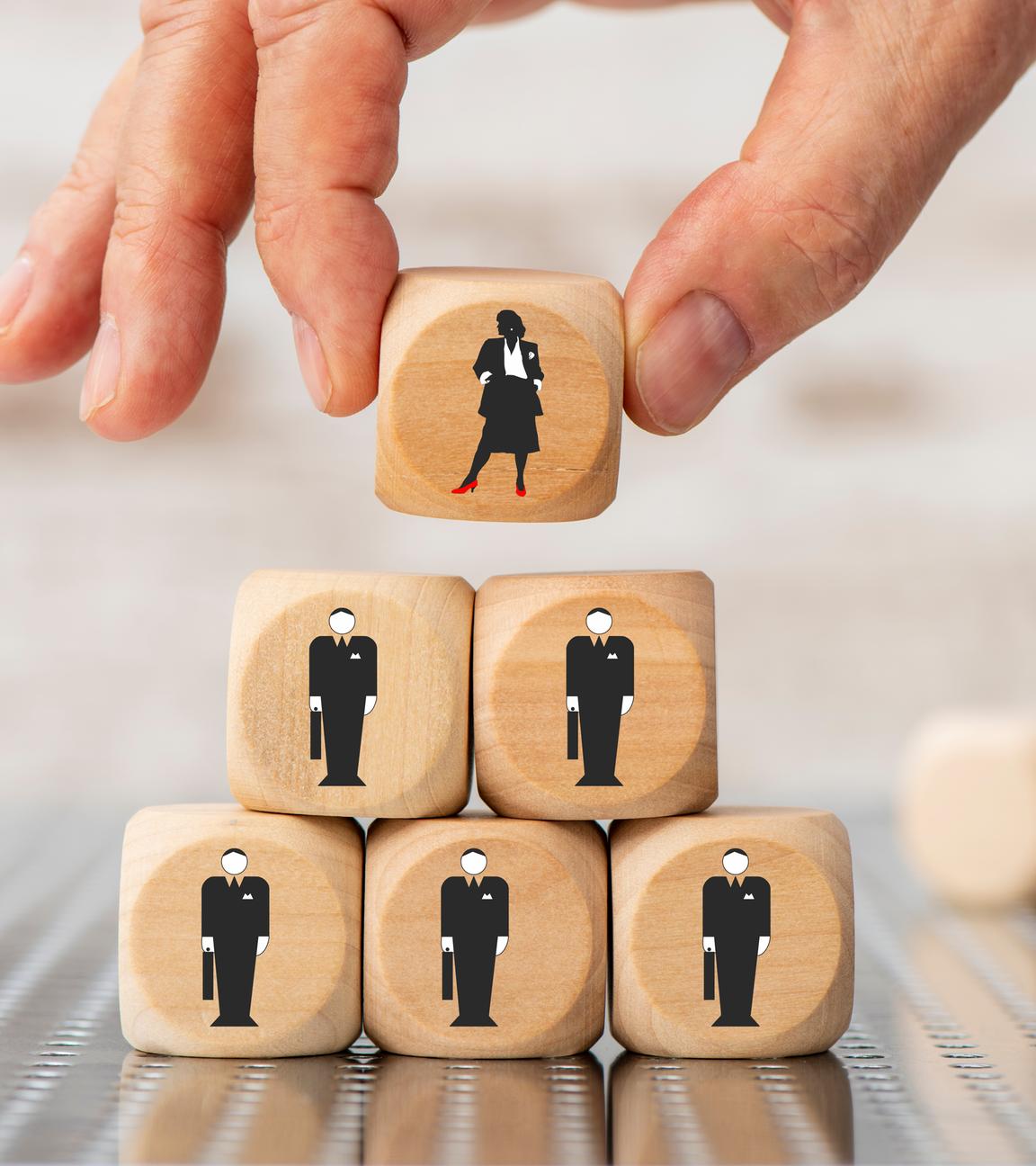 Eine Hand hält einen Würfel mit einem Frauen-Symbol. Darunter sind mehrere Würfel mit einem Männer-Symbol gestapelt.