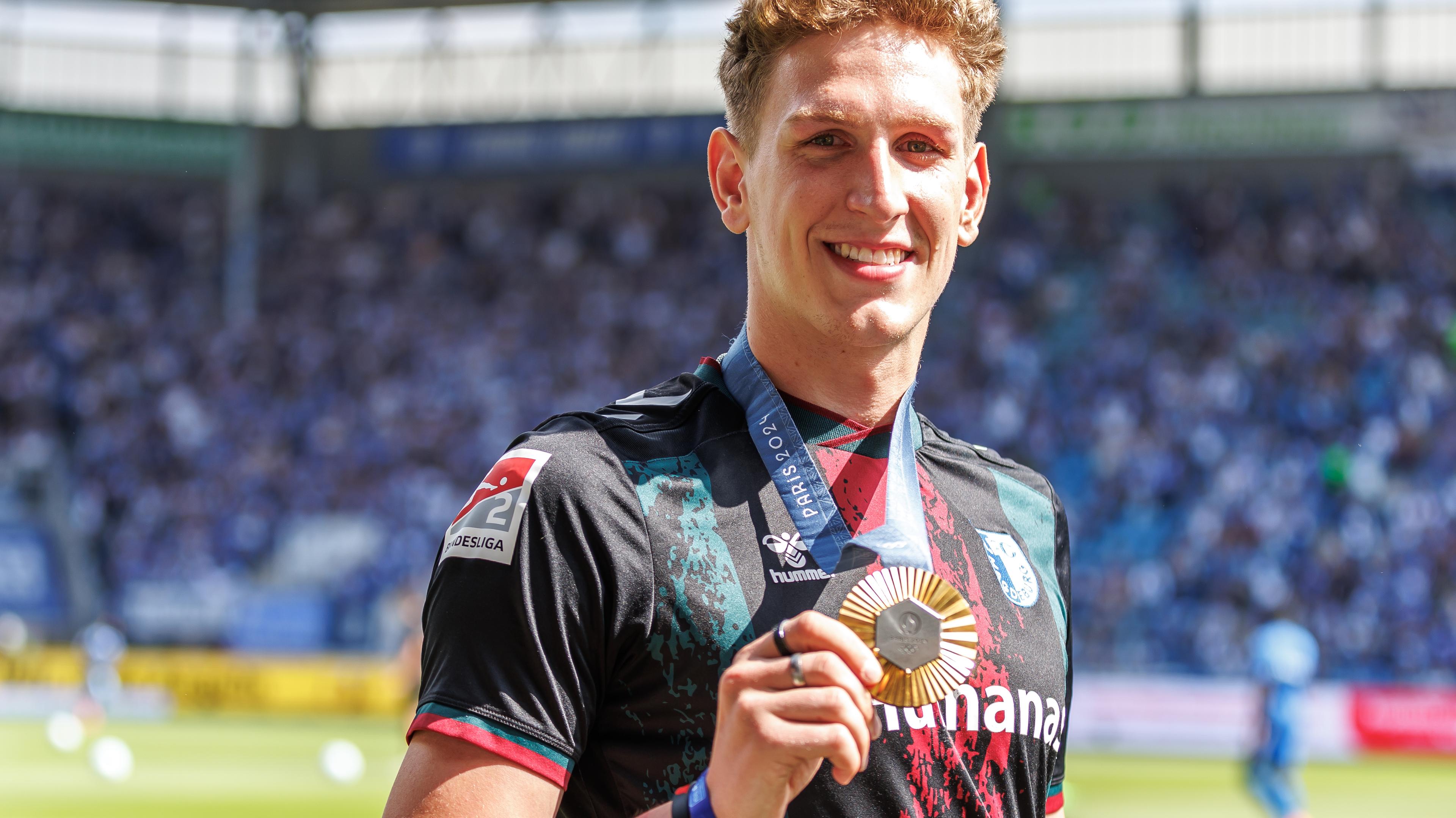 Schwimm-Olympiasieger Lukas Märtens strahlt mit seinem Olympia-Gold in einem signierten Trikot des 1. FC Magdeburg