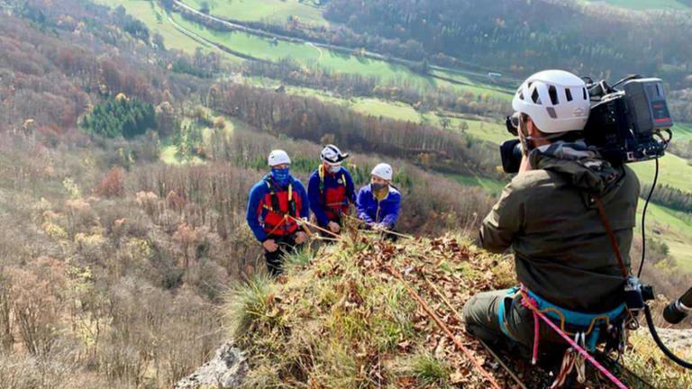 Auf dem Bild sind logo!-Reporterin Magdalena und zwei weitere Bergleute zu sehen, wie sie sich an einem Berghang abseilen. Im Vordergrund des Fotos kniet ein Kameramann, der die drei Personen filmt.