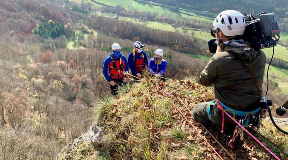 Auf dem Bild sind logo!-Reporterin Magdalena und zwei weitere Bergleute zu sehen, wie sie sich an einem Berghang abseilen. Im Vordergrund des Fotos kniet ein Kameramann, der die drei Personen filmt.