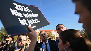 Ein Mann spricht in einer Menschenmenge in ein Mikrofon der Presse, während eine Person ein Schild mit der Aufschrift Nie wieder jetzt hoch hält