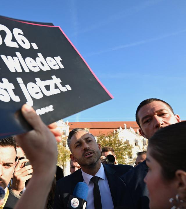 Ein Mann spricht in einer Menschenmenge in ein Mikrofon der Presse, während eine Person ein Schild mit der Aufschrift Nie wieder jetzt hoch hält
