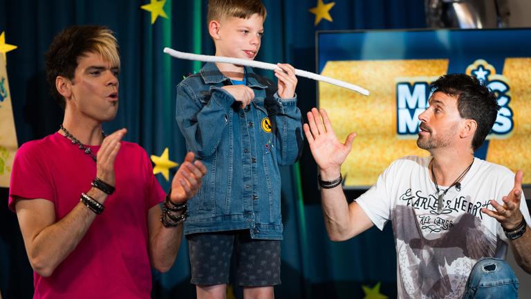 In einer Szene aus der ZDF-Doku "Magic Moves" stehen die Zauberer "Ehrlich Brothers" neben einem Kind, das gerade einen Zaubertrick vorführt.