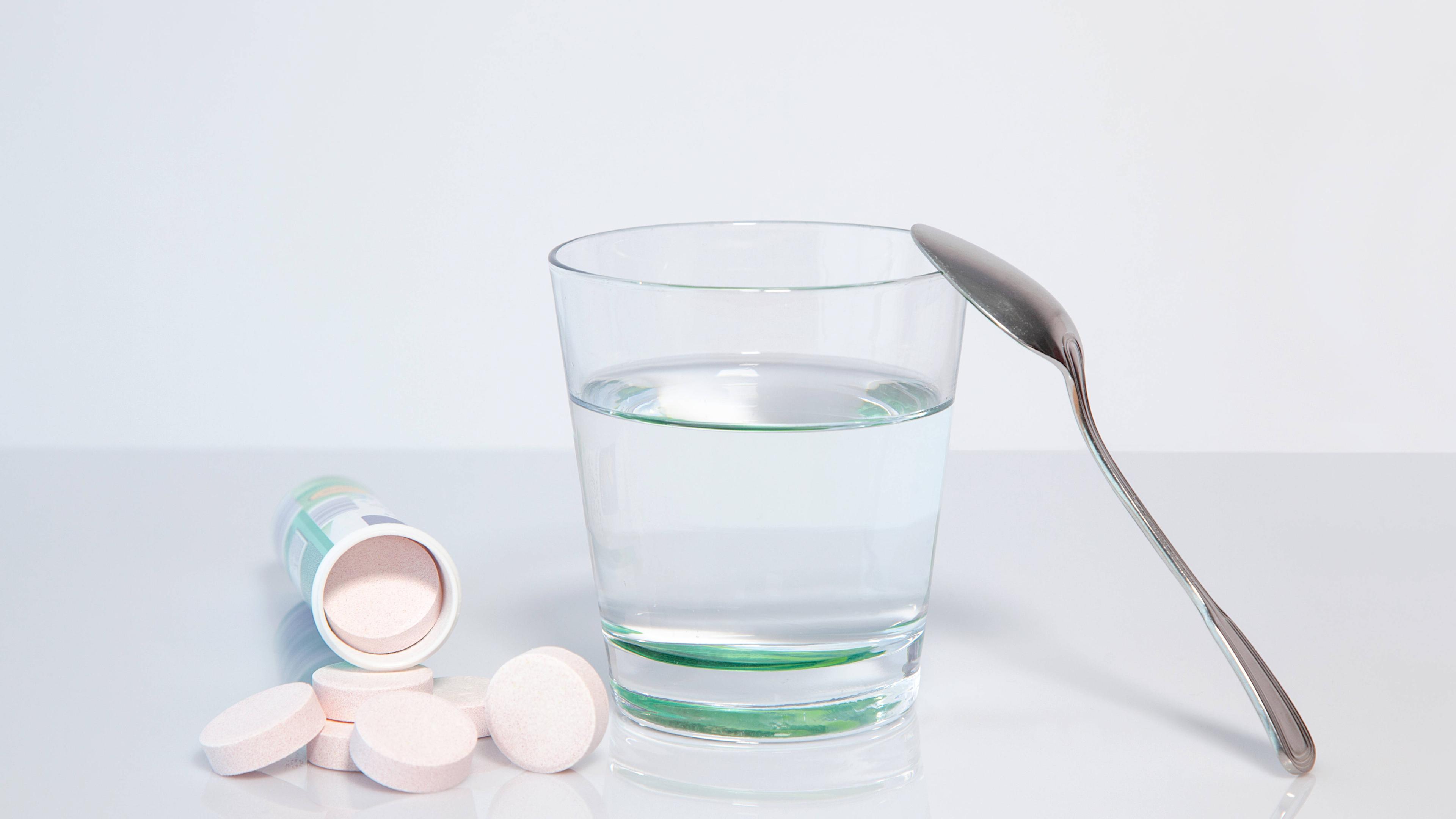 Mehrere Magnesium-Brausetabletten liegen vor einem umgekippten Röhrchen neben einem Wasserglas mit einem Löffel.