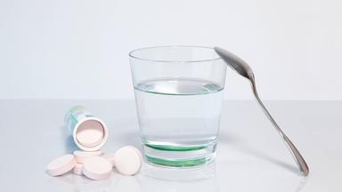 Mehrere Magnesium-Brausetabletten liegen vor einem umgekippten Röhrchen neben einem Wasserglas mit einem Löffel.