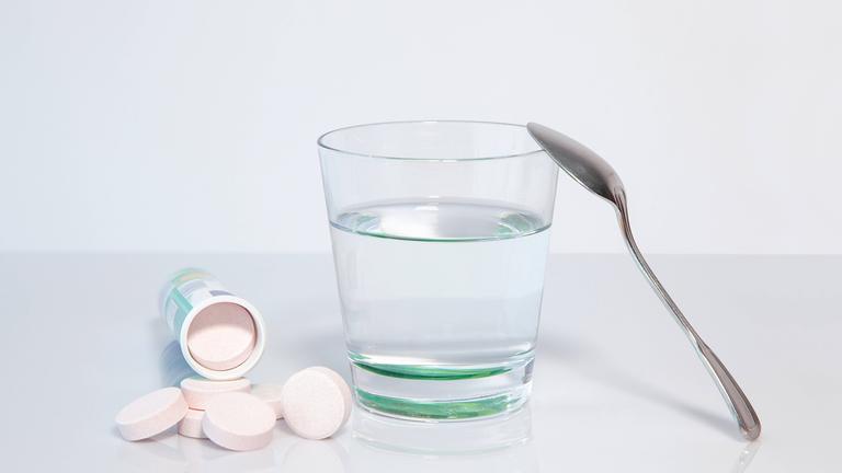 Mehrere Magnesium-Brausetabletten liegen vor einem umgekippten Röhrchen neben einem Wasserglas mit einem Löffel.