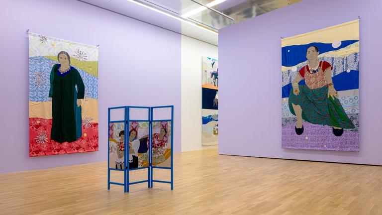 In einer Ausstellungshalle hängen zwei Gemälde, die jeweils eine Frau zeigen. Im Raum steht ein bemalter Paravent.