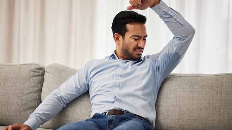 Ein Mann sitzt auf dem Sofa und betrachtet den Schweißfleck in der Achselhöhle auf seinem Hemd.