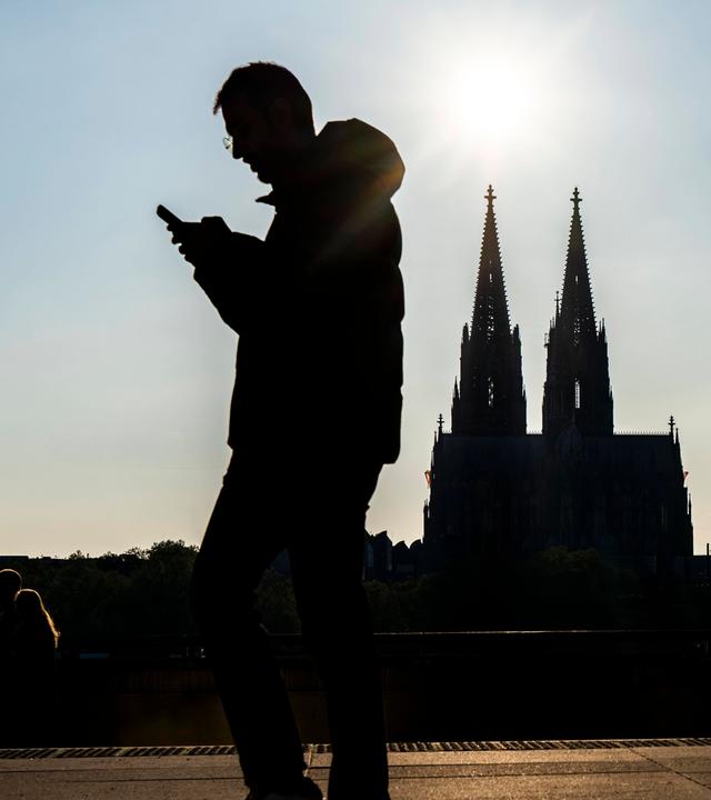 Silhouetten von einem Mann mit Handy in der Hand an der Rheinuferpromenade in Köln.