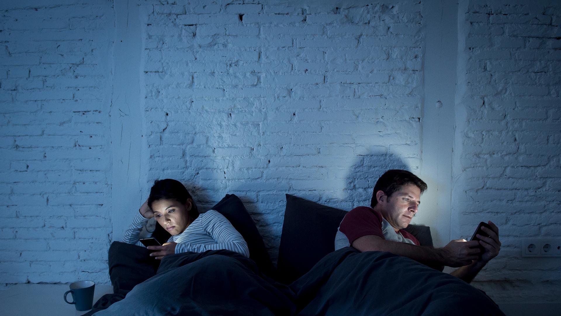 Ein Mann und eine Frau liegen abgewandt voneinander im Bett und schauen auf ihre Smartphones