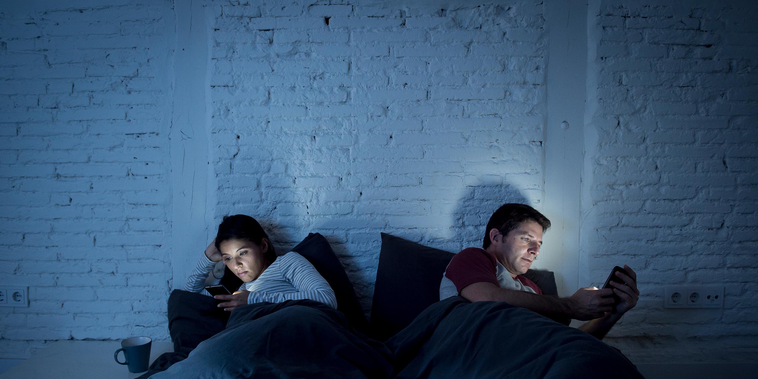 Ein Mann und eine Frau liegen abgewandt voneinander im Bett und schauen auf ihre Smartphones