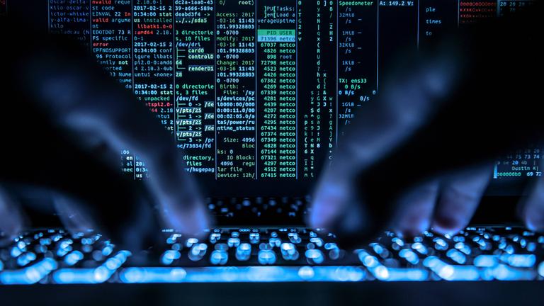 Ein Mann tippt auf der beleuchteten Tastatur eines Laptops. Auf dem Bildschirm ist ein Hackerprogramm geöffnet.