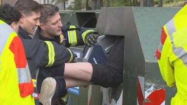 Auf dem Bild ist zu sehen: Zwei Feuerwehrleute beugen sich über die Öffnung eines grünen Elektrocontainers, in dem ein Mann feststeckt. Eine der Einsatzkräfte hält ein Werkzeug an der Einwurfklappe angesetzt.