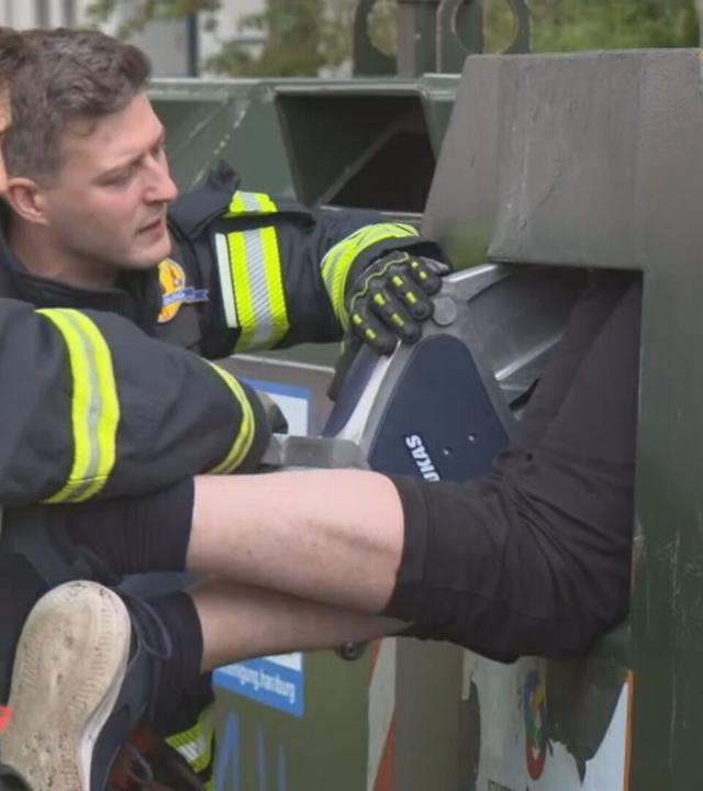 Auf dem Bild ist zu sehen: Zwei Feuerwehrleute beugen sich über die Öffnung eines grünen Elektrocontainers, in dem ein Mann feststeckt. Eine der Einsatzkräfte hält ein Werkzeug an der Einwurfklappe angesetzt.