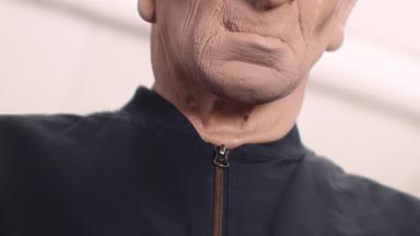 Person in schwarzer Jacke mit menschenähnlicher Maske, deren Gesicht nur teilweise zu sehen ist.