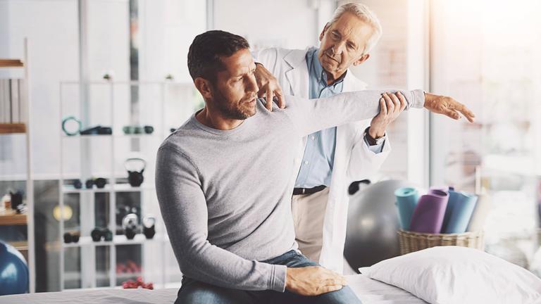 Ein Mann mit Schmerzen in der Schulter beim Anheben des Arms bei er Untersuchung durch einen Arzt.