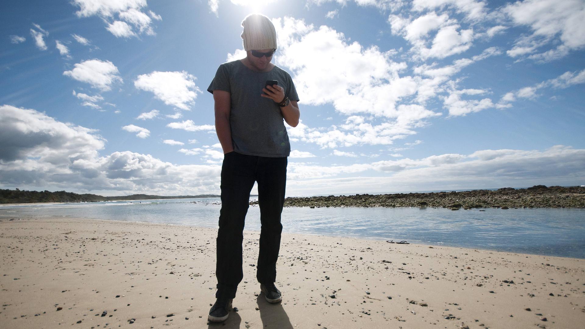 Mann steht am australischen Strand und schaut auf sein Handy 
