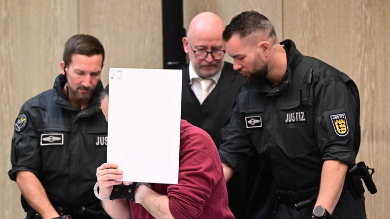 Der Angeklagte in einem Mordprozess nach einer Todesfahrt von Mannheim sitzt mit einem Aktendeckel vor seinem Gesicht im Gerichtssaal, rechts neben ihm steht sein Verteidiger Uwe Kosmala.