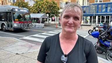 Eine ältere Dame mit Kurzharrschnitt seht am Rande einer Straße.