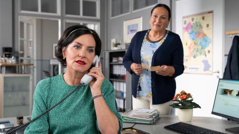 Miriam Stockl (Marisa Burger) sitzt am Schreibtisch ihres Büros und telefoniert mit ernster Miene. Sie hat Marianne Grasegger (Ursula Maria Burkhart), die hinter ihr steht und sie ansieht, den Rücken zugewandt.