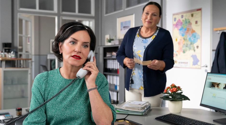 Miriam Stockl (Marisa Burger) sitzt am Schreibtisch ihres Büros und telefoniert mit ernster Miene. Sie hat Marianne Grasegger (Ursula Maria Burkhart), die hinter ihr steht und sie ansieht, den Rücken zugewandt.
