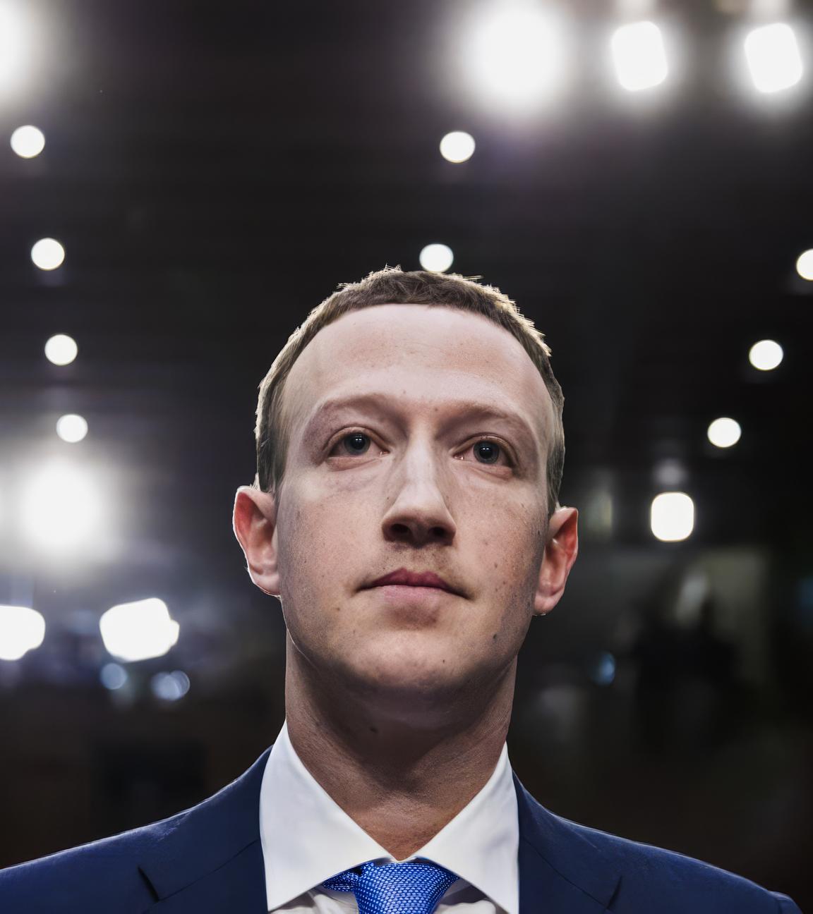 Frontale Aufnahme von Mark Zuckerberg in Anzug und Krawatte in einer Anhörungssituation, an der Decke sind zahlreiche Scheinwerfer, im Hintergrund ist verschwommen eine Menschenmenge zu erkennen.