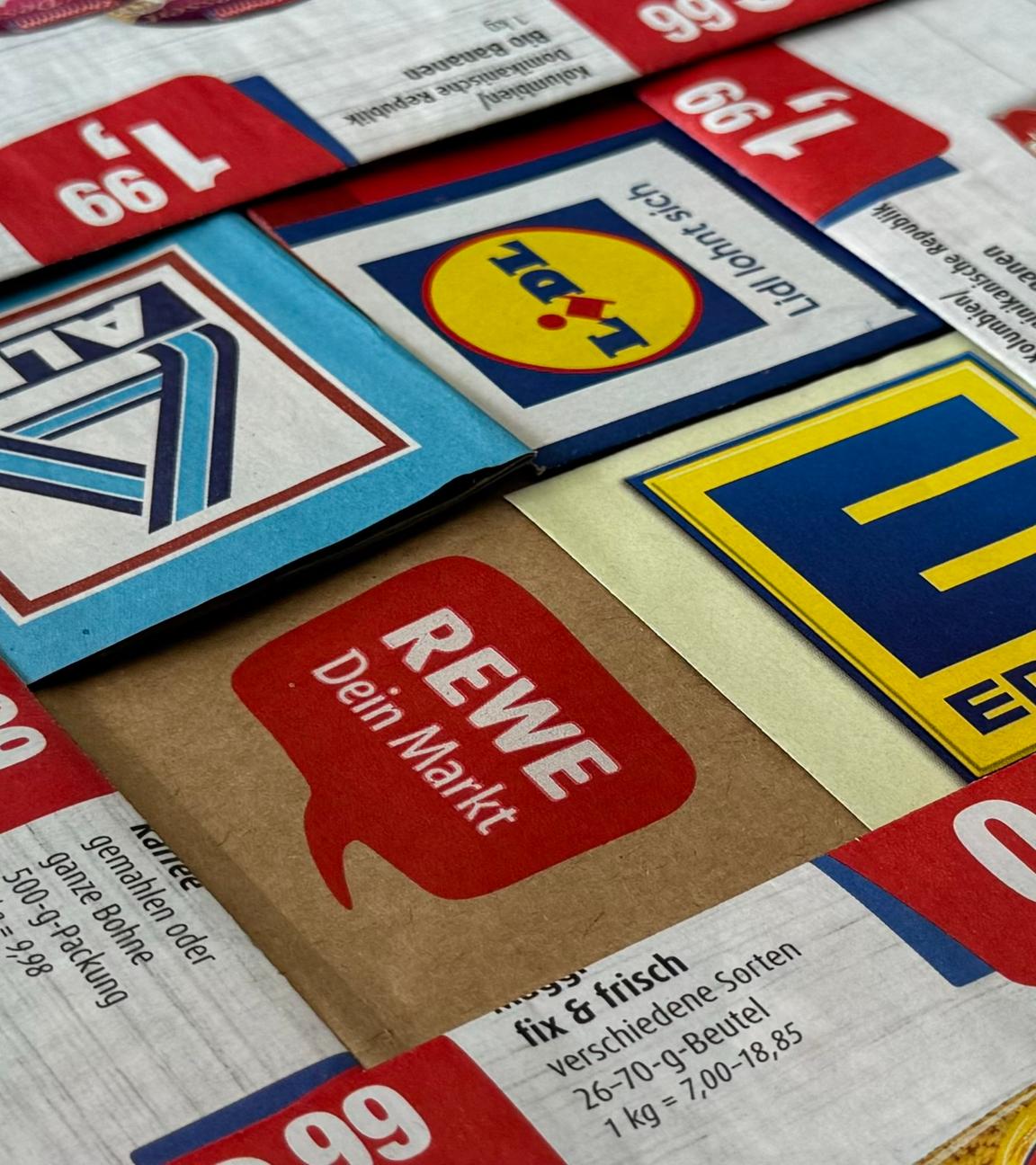 Logos und Prospekte von Edeka, Rewe, Aldi und Lidl liegen eng beisammen.