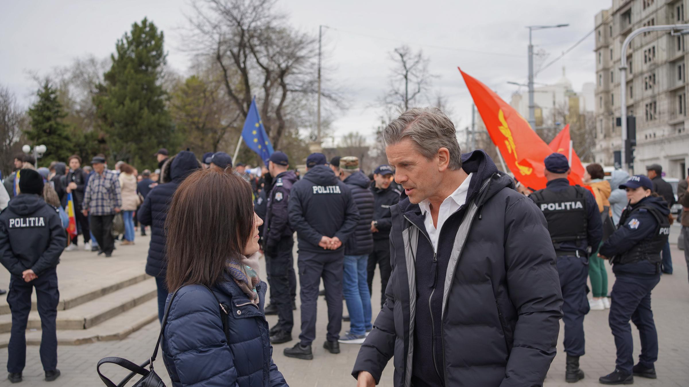 "Markus Lanz – Moldawien ungeschminkt": Markus Lanz und eine Frau stehen auf einer Straße und unterhalten sich. Im Hintergrund ist eine Menschenmenge zu sehen, an deren Rand Polizisten stehen.