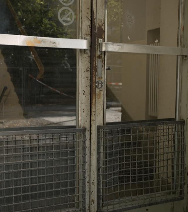 Vergitterte, verwitterte Glastür mit Metallquerstreben und Rostspuren in einem hellen, verschmutzten Uni-Gebäudeeingang.