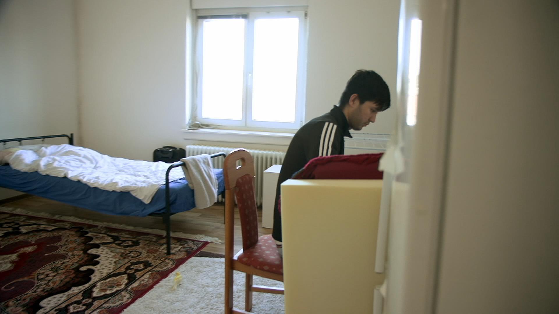 Mann sitzt in seinem Zimmer am Schreibtisch, im Hintergrund steht ein Bett