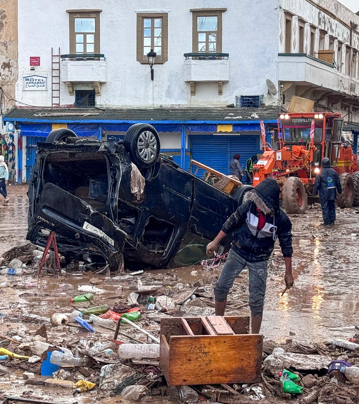 Menschen in einer verwüsteten Straße voller Schlamm. Ein Auto liegt auf dem Dach.