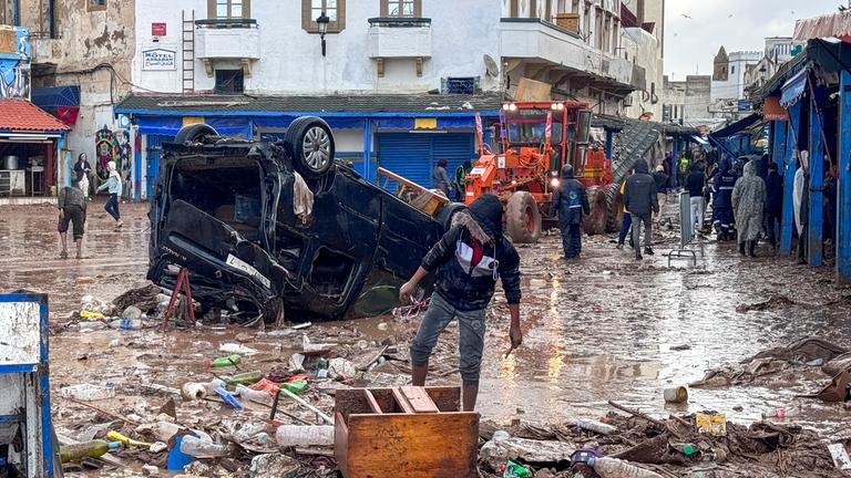 Menschen in einer verwüsteten Straße voller Schlamm. Ein Auto liegt auf dem Dach.