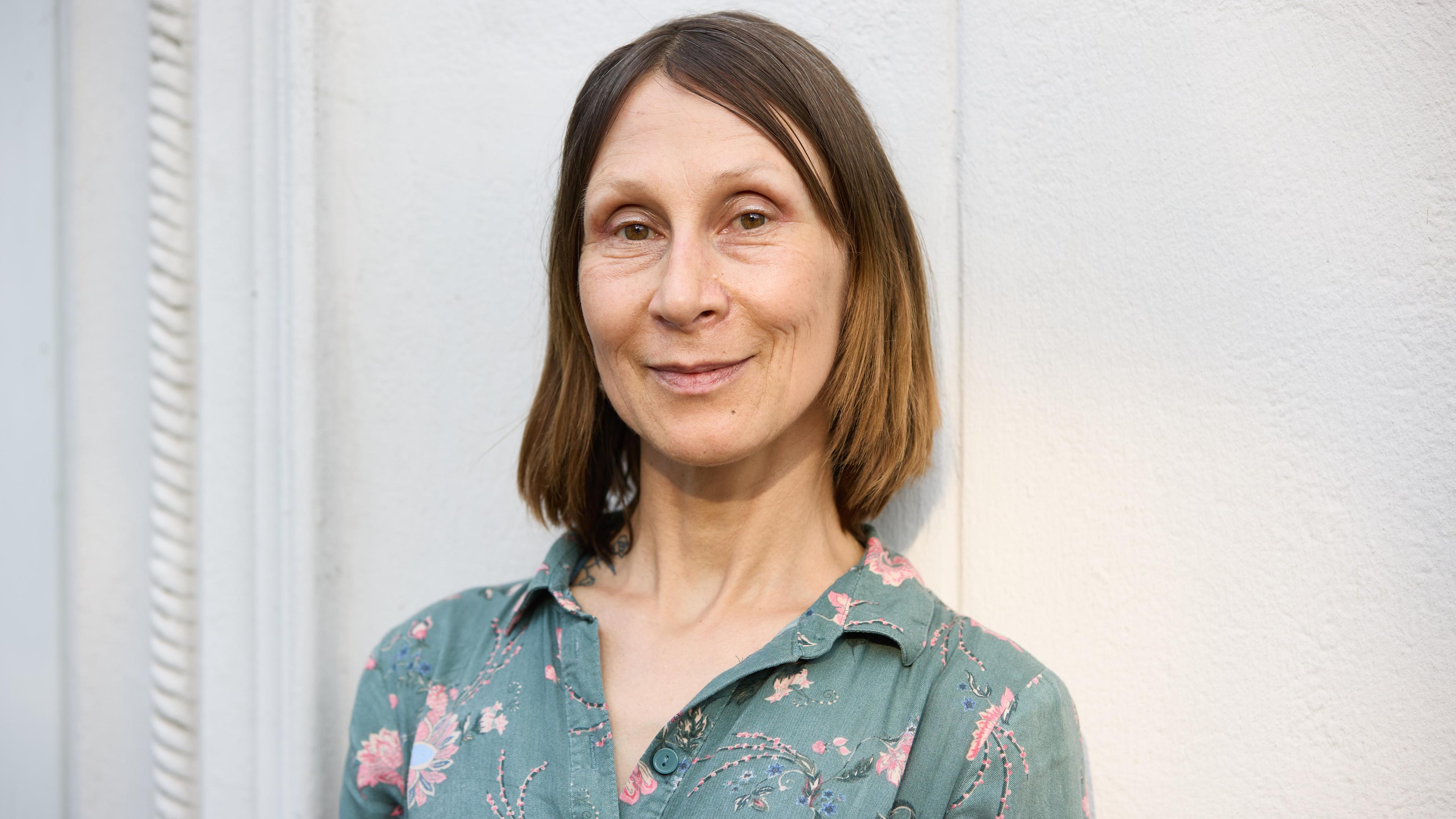 Eine Frau mittleren Alters mit schulterlangen glatten mittelblonden Haaren trägt eine grüne Bluse mit rosa Blumen und schaut lächelnd in die Kamera.