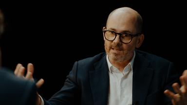 Mann in seinen frühen 50ern mit sehr kurzen grauen Haaren und gestutztem bronzenen Bart sitzt in einem braunen Sessel vor einem schwarzen Hintergrund. Er trägt eine schwarze Brille, ein dunkles Jackett und ein weißes Hemd.