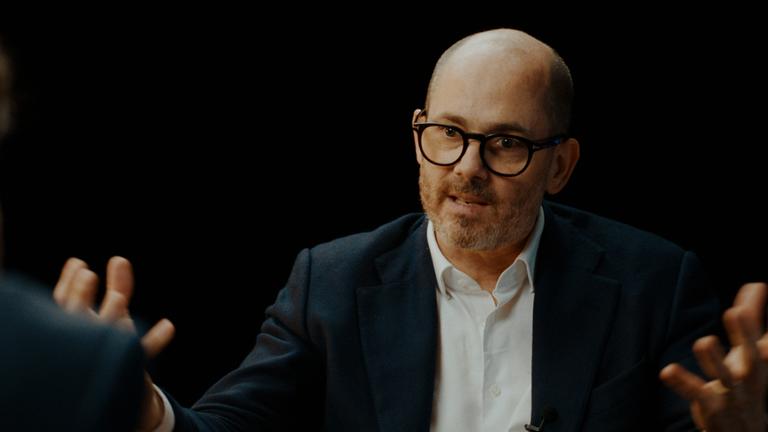 Mann in seinen frühen 50ern mit sehr kurzen grauen Haaren und gestutztem bronzenen Bart sitzt in einem braunen Sessel vor einem schwarzen Hintergrund. Er trägt eine schwarze Brille, ein dunkles Jackett und ein weißes Hemd.