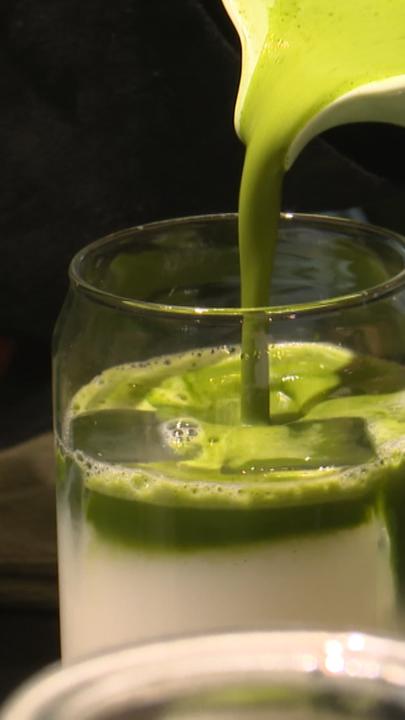 Ein Glas Milch, in das mit einem Kännchen grüner Matcha-Tee gegossen wird.