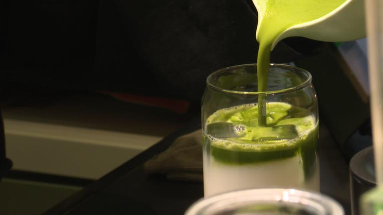 Ein Glas Milch, in das mit einem Kännchen grüner Matcha-Tee gegossen wird.