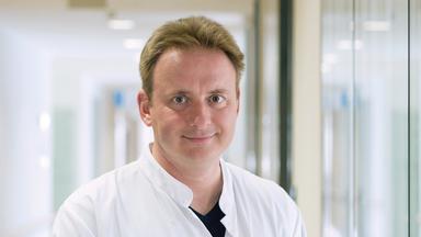 Professor Matthias Blüher steht im Gang einer Klinik und lächelt in die Kamera.