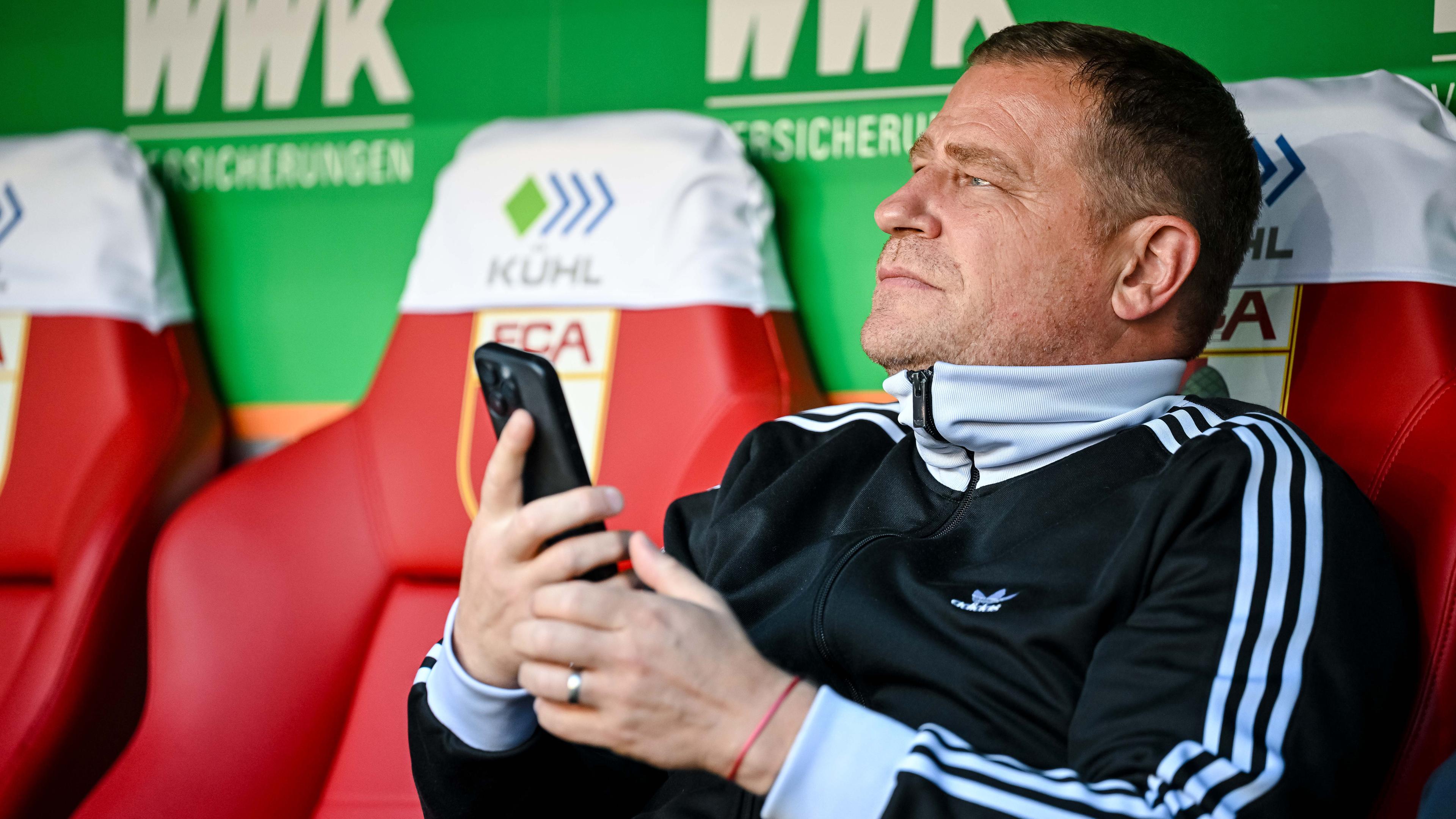 Max Eberl, Sportvorstand FC Bayern München, sitzt vor dem Spiel mit dem Handy auf der Bank.