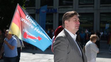 Ein Mann in Anzug läuft auf der Straße, im Hintergrund trägt ein Mann eine Flagge, auf der Unser Land, unsere Heimat und Alternative für Deutschland steht.