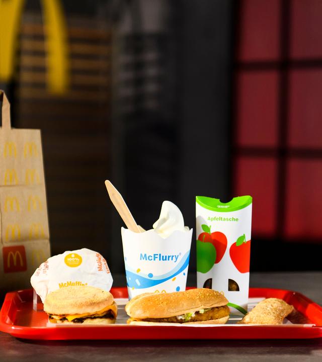 Auf einem Tablett sind ein McChicken, ein McFlurry, ein McMuffin und eine Apfeltasche drapiert. Daneben steht eine McDonald's-Tragetasche.