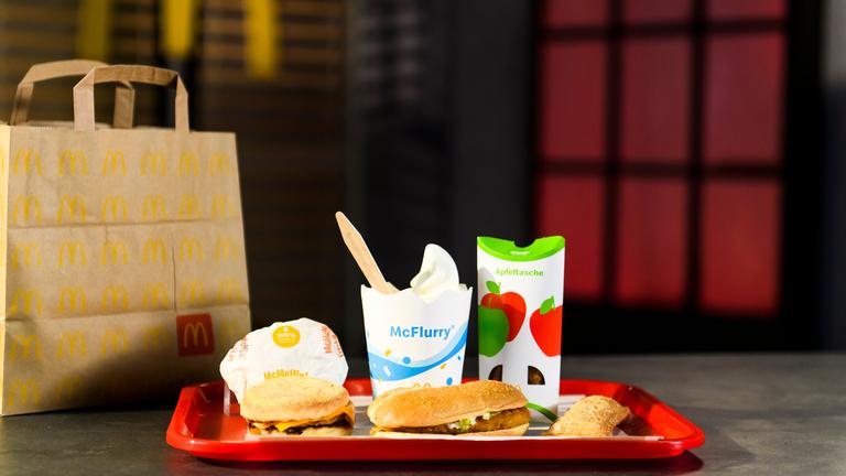 Auf einem Tablett sind ein McChicken, ein McFlurry, ein McMuffin und eine Apfeltasche drapiert. Daneben steht eine McDonald's-Tragetasche.