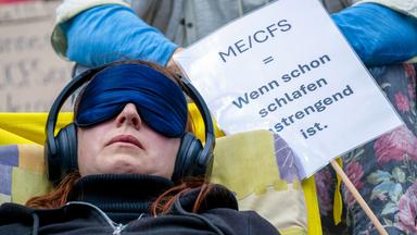 Eine Frau liegt mit einer Schlafmaske und Kopfhörern auf einer Liege neben einem Schild mit der Aufschrift «ME/CFS = Wenn schon schlafen anstrengend ist.»
