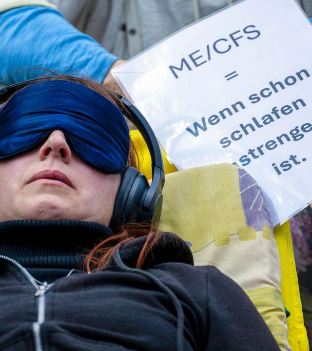 Eine Frau liegt mit einer Schlafmaske und Kopfhörern auf einer Liege neben einem Schild mit der Aufschrift «ME/CFS = Wenn schon schlafen anstrengend ist.»