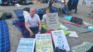 Menschen liegen und sitzen auf Kopfsteinpflaster, sie halten verschiedene Plakate.
