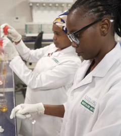 Zwei dunkelhäutige Mitarbeiterinnen in weißem Kittel arbeiten in einem Labor.