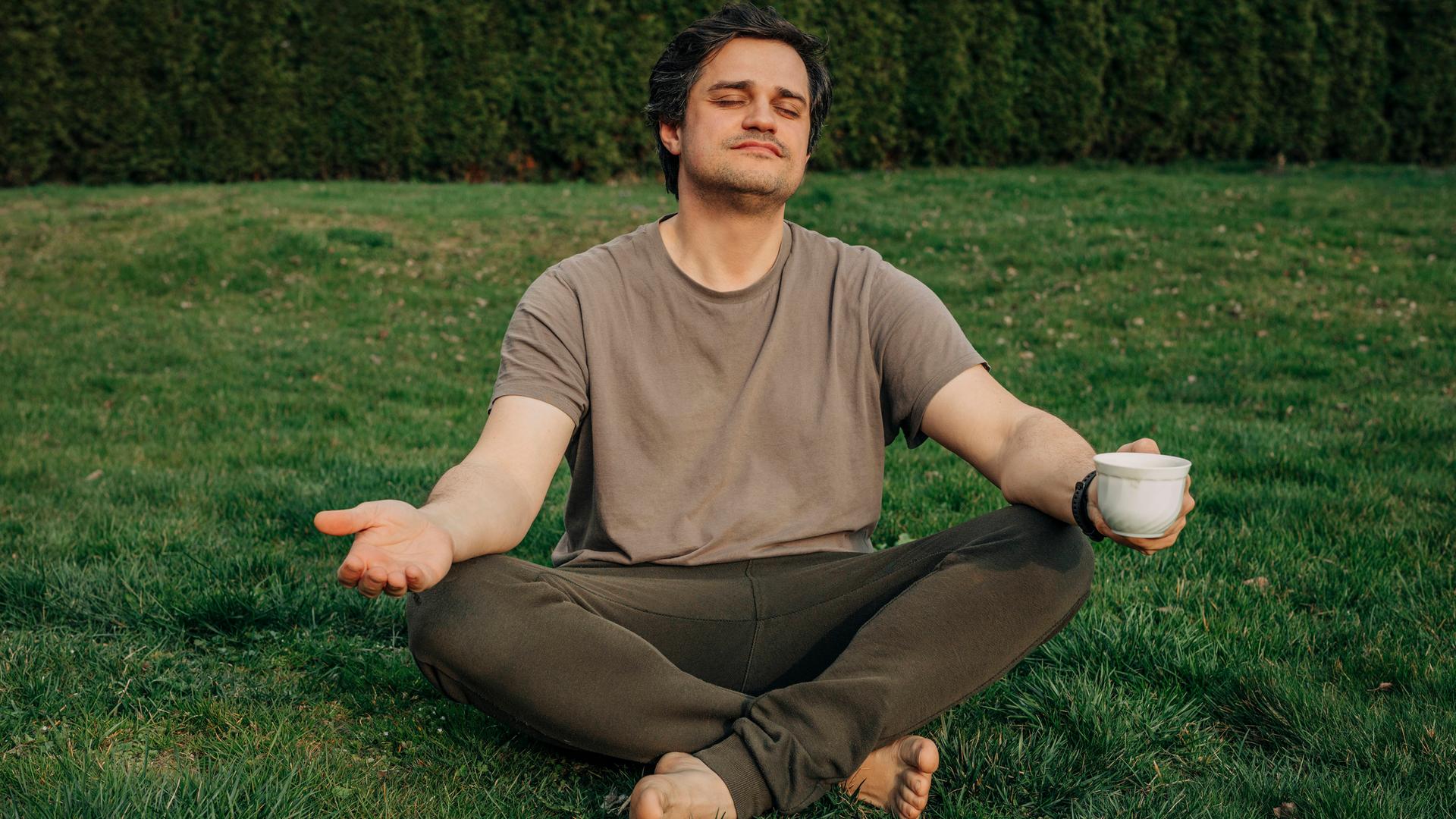 Mann im Schneidersitz mit Kaffeetasse in der Hand meditiert im Garten. 