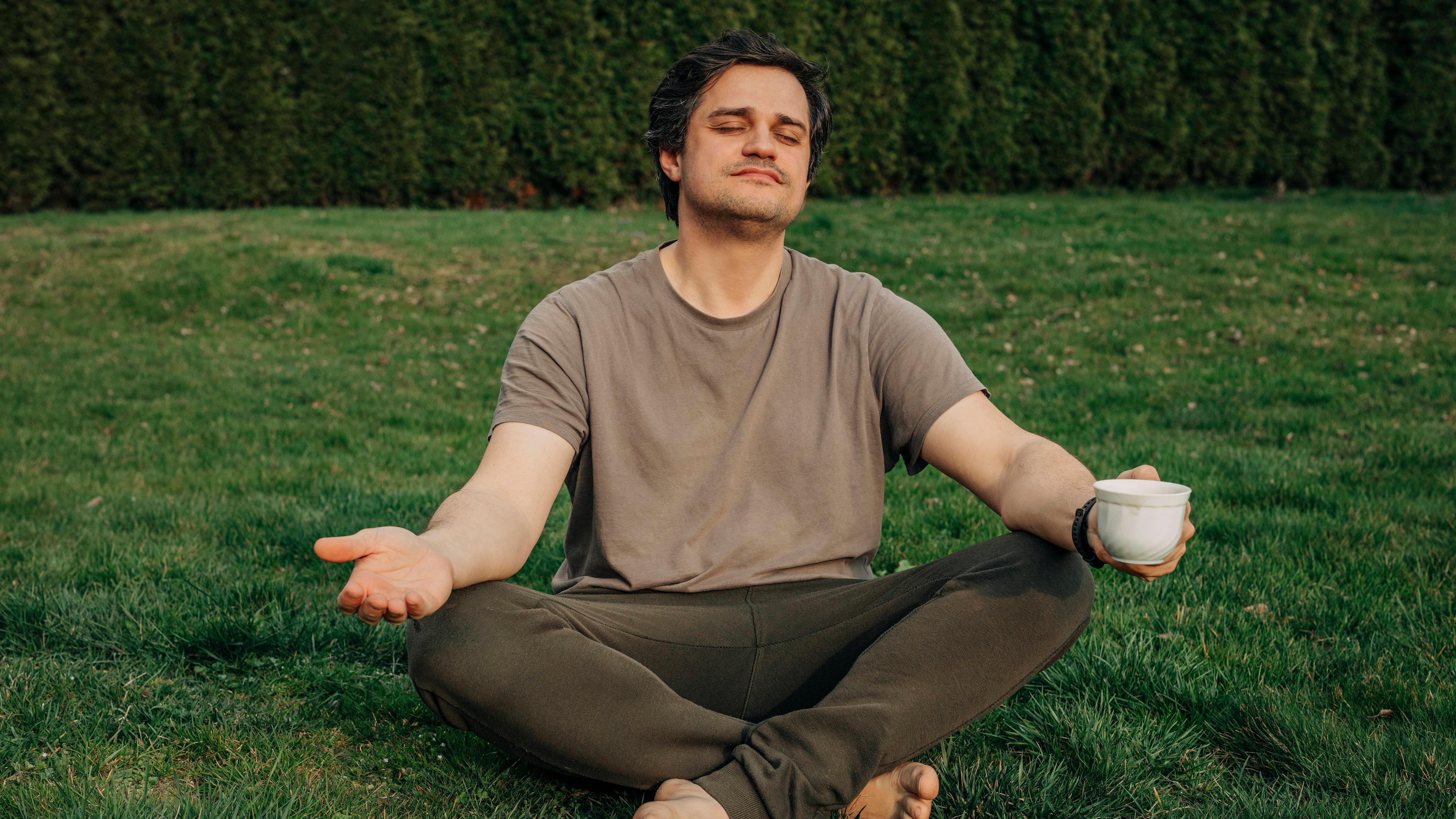 Mann im Schneidersitz mit Kaffeetasse in der Hand meditiert im Garten. 