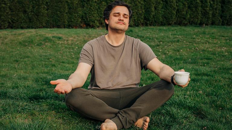 Mann im Schneidersitz mit Kaffeetasse in der Hand meditiert im Garten. 