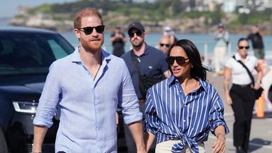 Auf dem Bild zu sehen ist: Meghan und Harry Hand in Hand eine Straße entlang laufen. Die Perspektive ist eine Halbtotale.
